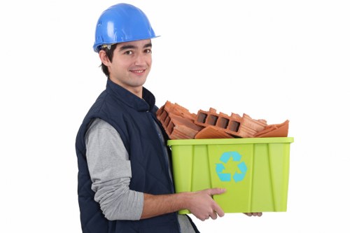 A young man wearing a blue hard hat and a navy work jacket with grey sleeves is standing against a plain white background, holding a bright green recycling bin marked with the recycling symbol. Inside the bin, there are multiple red clay bricks stacked neatly, with some bricks leaning against the side. The man is smiling slightly and pointing towards the bricks with his right hand, presenting the contents of the bin. The overall scene appears to be set in a professional context related to waste or rubbish removal, with a focus on construction debris recycling. The plain white backdrop ensures the subject and the bin are clearly visible, emphasizing the company's rubbish removal services in areas such as Covent Garden or nearby London neighborhoods.