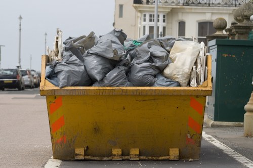 A large yellow skip bin situated on a street in a residential or commercial area, filled with black and white rubbish bags made of plastic and cloth materials. The skip shows signs of dirt and wear on its exterior, with reflective orange safety markings on the sides. Behind the skip, a row of parked cars and a glimpse of a white building with decorative architectural features are visible, indicating an urban environment potentially within Covent Garden or nearby London postcode. The street surface appears to be asphalt, with another green waste collection container partly visible to the right. The image reflects a typical rubbish clearance scene, emphasizing waste removal services such as Covent Garden House Clearance, operating in local areas, and highlights the process of collecting household or commercial waste for disposal or recycling.