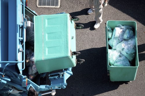 An overhead view of a green waste collection vehicle with its rear compartment open, showing a large bin filled with clear plastic bags containing mixed household rubbish. Next to the vehicle, a green wheeled bin is positioned on the dark asphalt surface, also filled with sorted waste, including plastic packaging and other refuse. Partially visible, a person’s legs in light-colored clothing and white footwear are standing nearby, indicating active rubbish collection or disposal. The scene takes place outdoors, possibly on a street or driveway, with the focus on the practical task of rubbish removal in an urban or residential area, typical of a service operated by Covent Garden House Clearance in London.