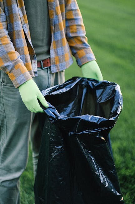 A man wearing a black cap, red t-shirt, and light blue shorts is standing on a sandy beach, holding a white plastic bag filled with collected rubbish. To his left, a young boy in a grey t-shirt, beige