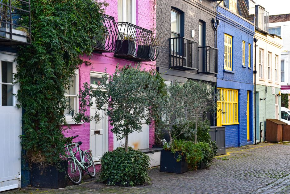 A row of traditional Victorian terraced houses painted in varying shades of blue, featuring bay windows, decorative cornices, and brick steps leading to small front gardens. The buildings' facades dis