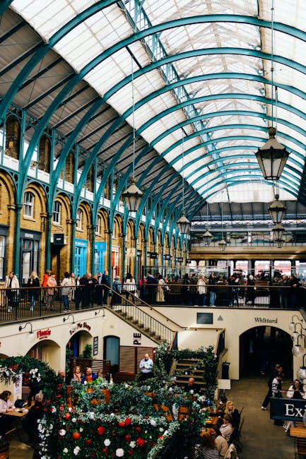 Inside a large indoor marketplace with a high, arched glass ceiling supported by blue-tinted metal framework, the scene shows a busy public space filled with people walking and standing throughout. On
